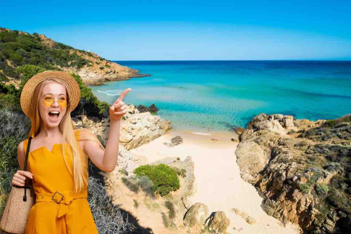 Donna vestita di giallo con occhiali e cappello indica con il doto e sul retro spiaggia sabbiosa e con rocce