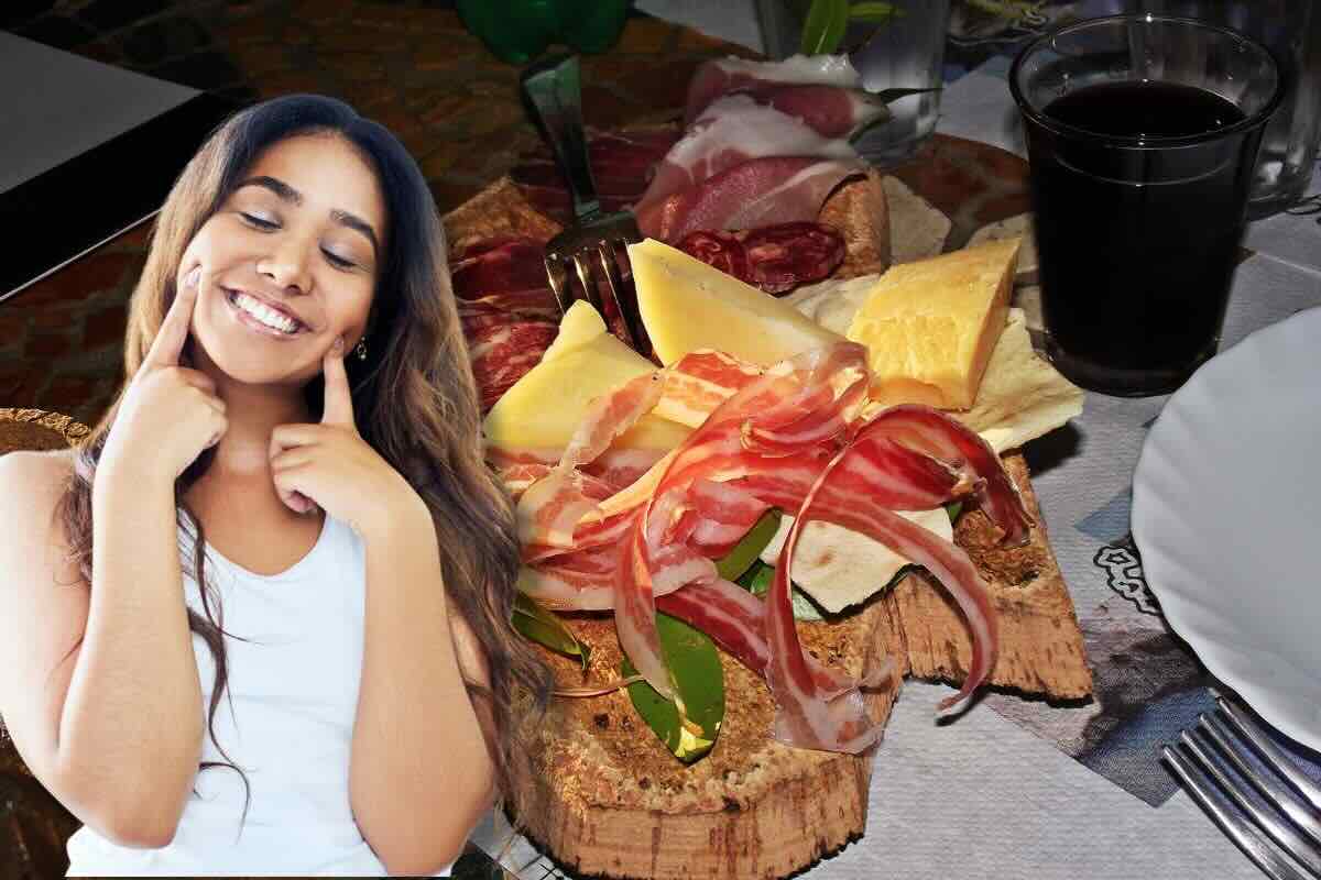 Ragazza con dita sulle guance, sorridente, dietro di lei tagliere salumi e formaggi e vino